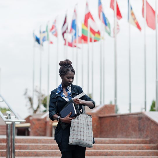 African student female posed with backpack and school items on yard of university, against flags of different countries.